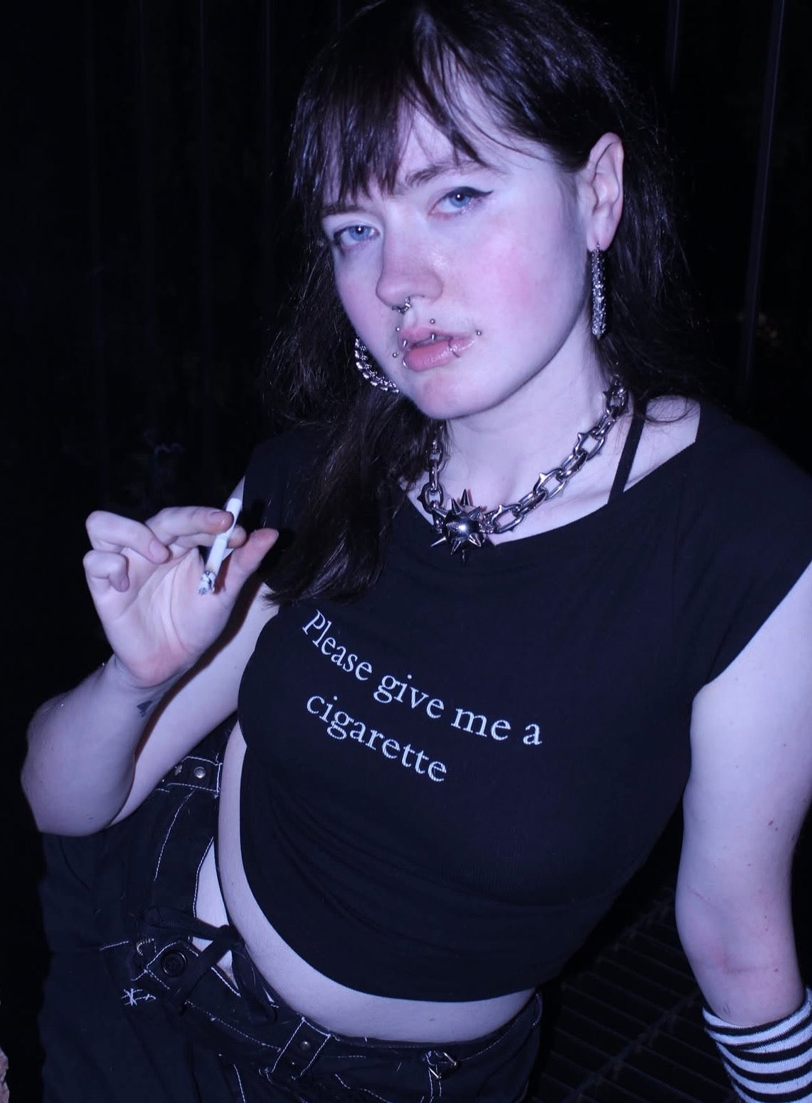 Person wearing a black tank top with text, holding a cigarette against a dark background