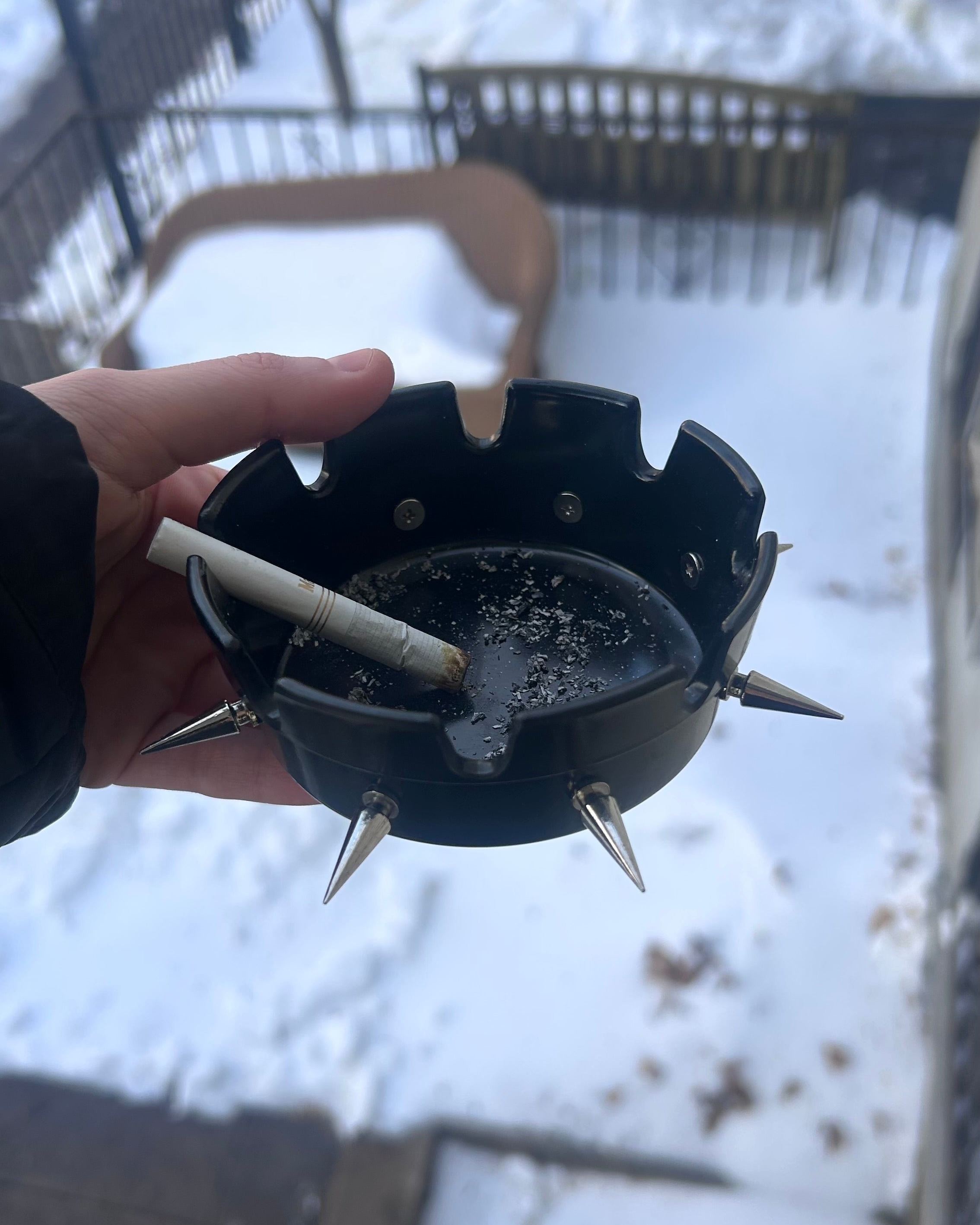 Hand holding a black ashtray with spikes in front of a snowy background 