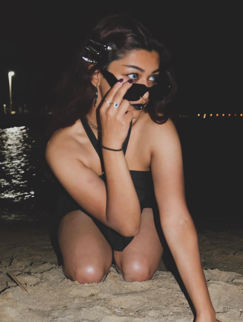 A woman in a black one-piece swimsuit kneels on sand at night near the water, wearing black sunglasses and looking toward the camera.