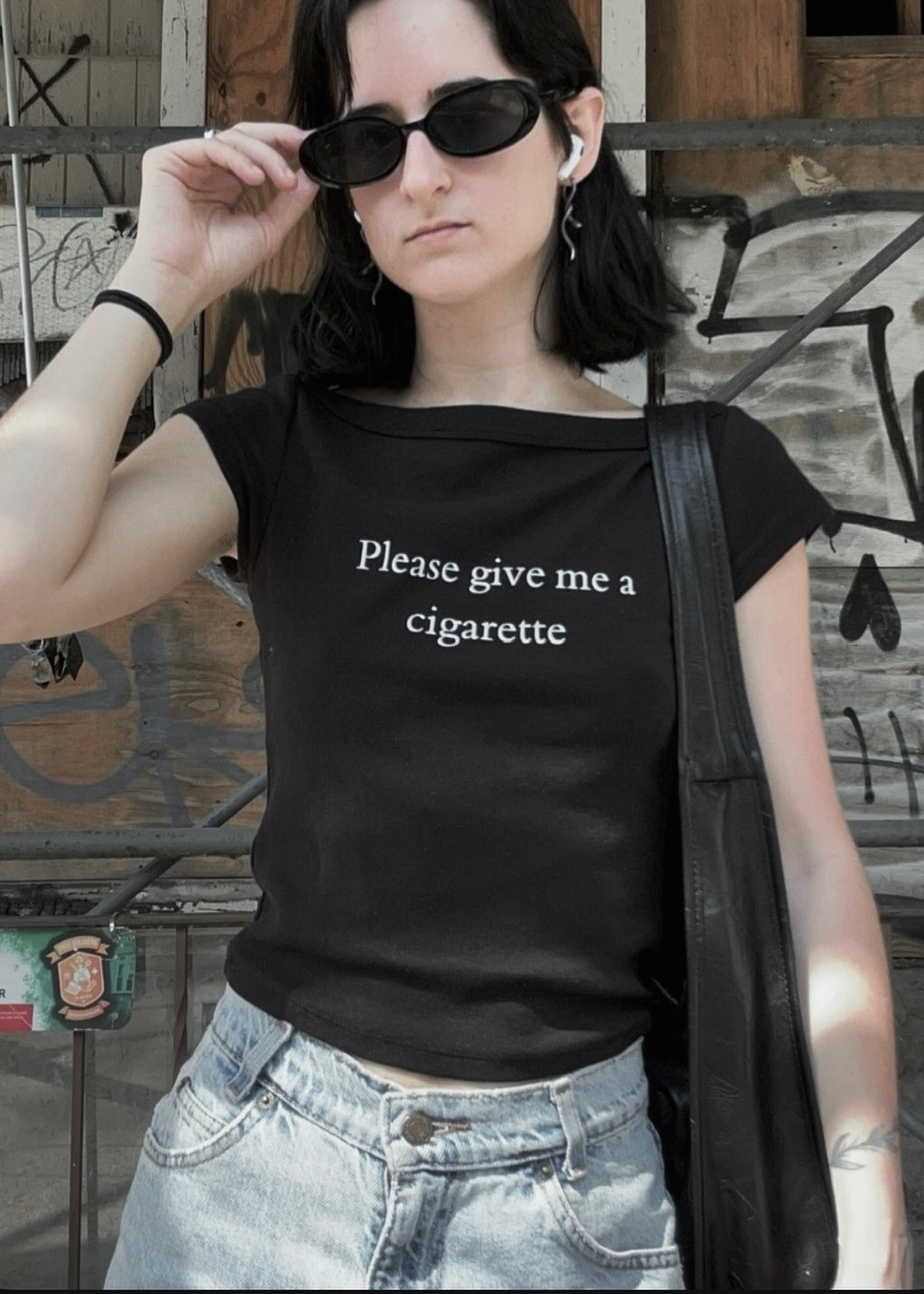 A woman wearing a black off-the-shoulder t-shirt with the white text 'Please give me a cigarette' printed on the front, paired with light-wash denim jeans and black sunglasses, standing against a graffiti-covered wooden wall.
