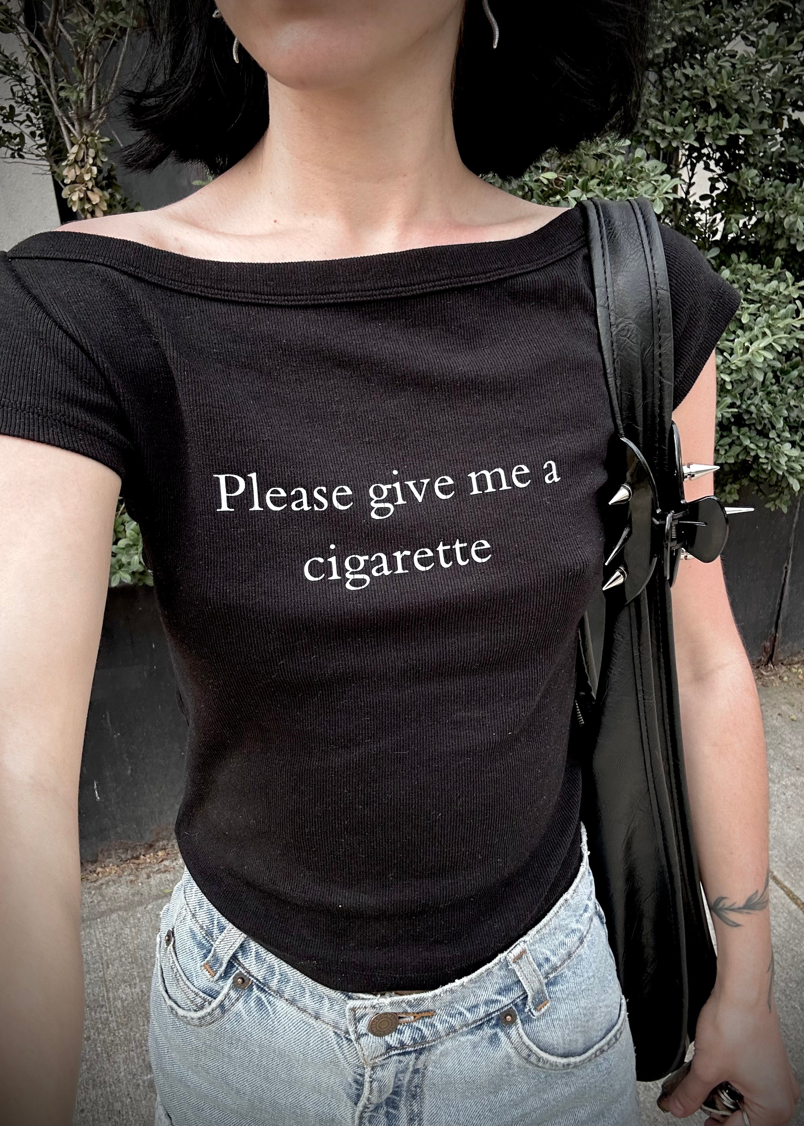 Person wearing a black t-shirt with 'Please give me a cigarette' text outdoors.