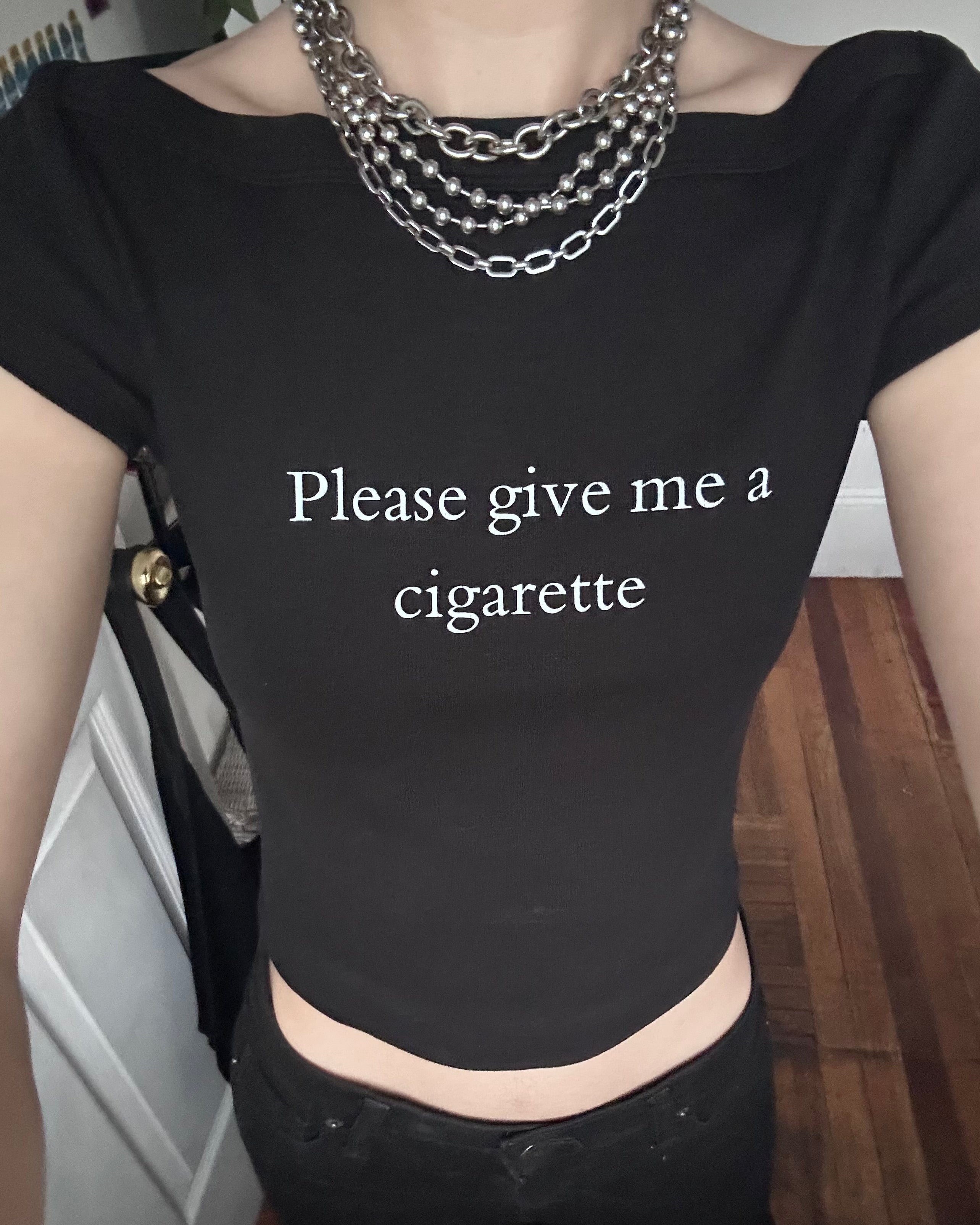 Person wearing a black t-shirt with 'Please give me a cigarette' text, standing indoors.
