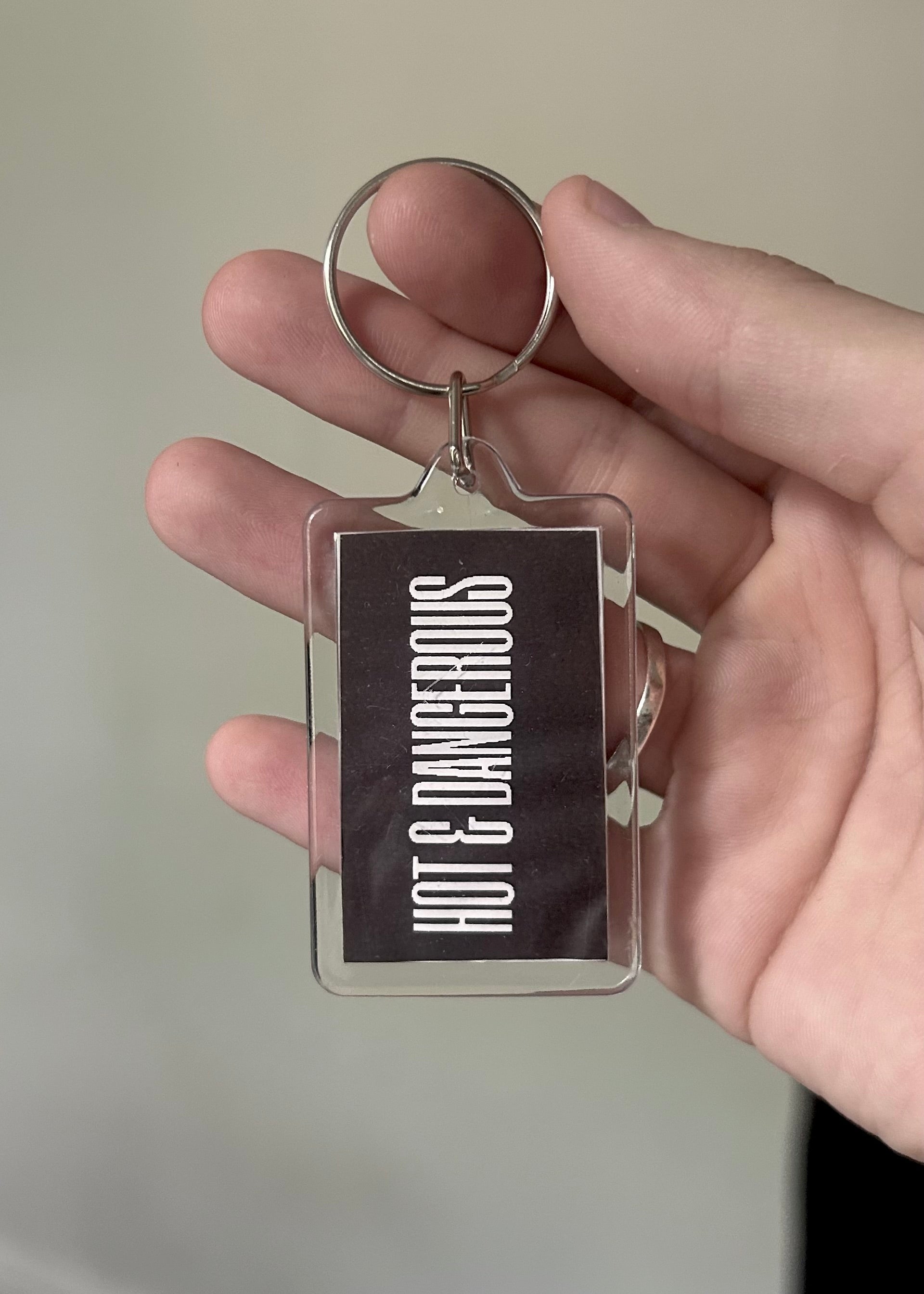 Keychain with 'Hot & Dangerous' text held in a hand against a neutral background