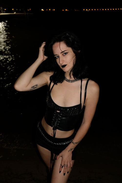 A woman posing outdoors at night wearing a black strappy corset-style top and matching shorts with silver chain accents.