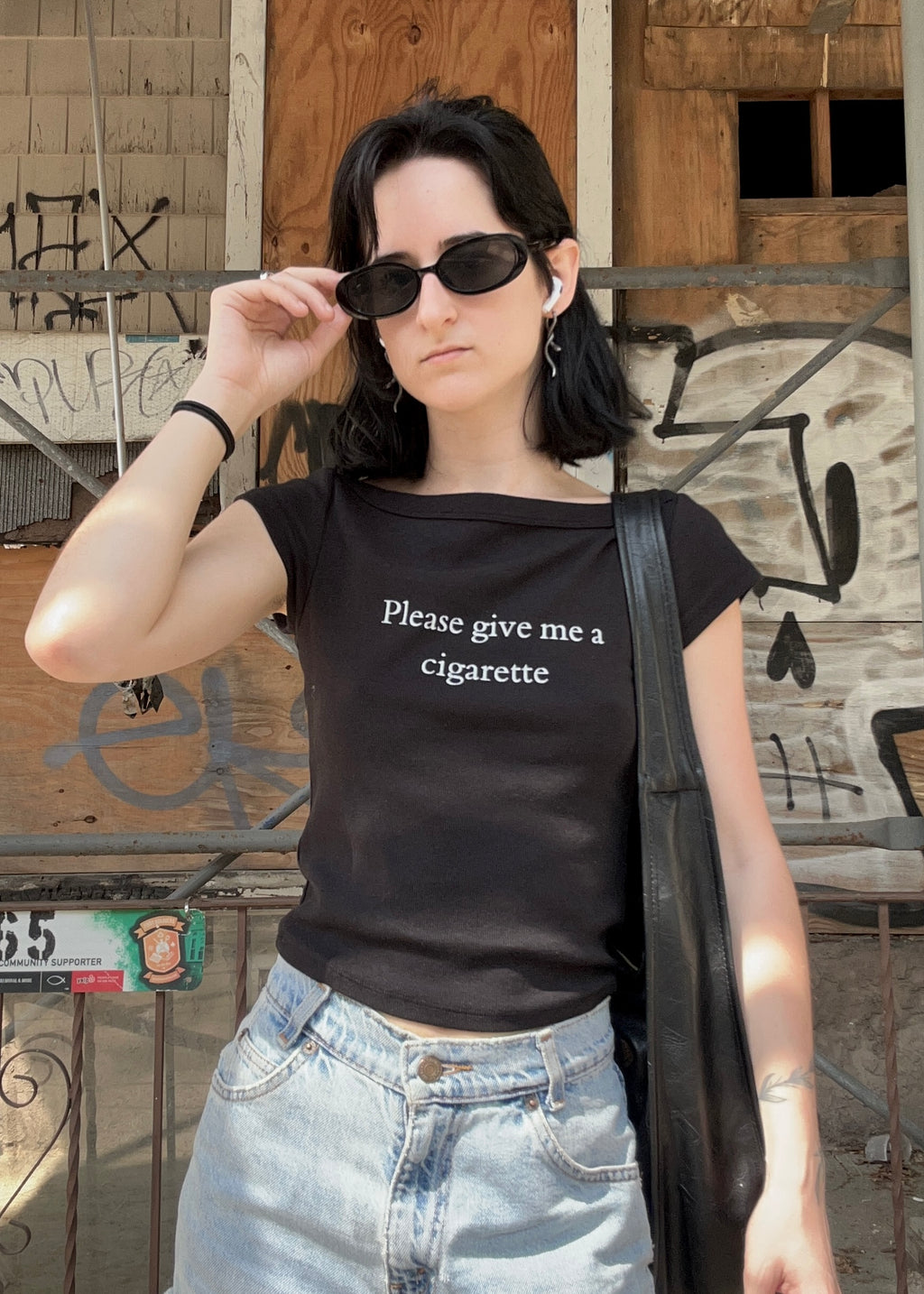 Person wearing a black t-shirt with text, standing in front of a graffiti-covered wall.